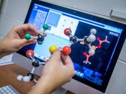 Hands holding models of chemical bonds in front of a computer display screen.