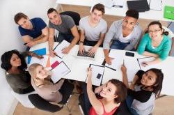 a group is seated around a table with paper and laptops working together. camera is above and they are turning their heads up towards the camera.