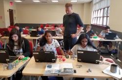 Professor observes work done by high school students who are sitting at laptops