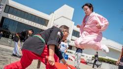 A female student in pink jumps high over a paddle held by a man in red and black as a woman looks on.