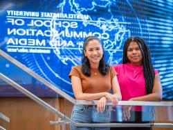 Two students stand before a Montclair State University School of Communication and Media screen.