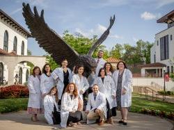 Eleven students wearing white lab coats pose for a photo in front of a bronze red hawk statue.