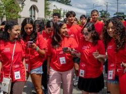 A group of students in red t-shirts and lanyards walk together in a large group on college campus.