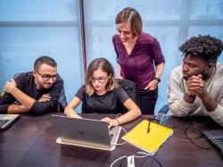 Three students sitting at table looking at a laptop while professor looks on from behind.