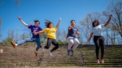 Four students wearing different t-shirts representing their College or School jumping in the University Amphitheater